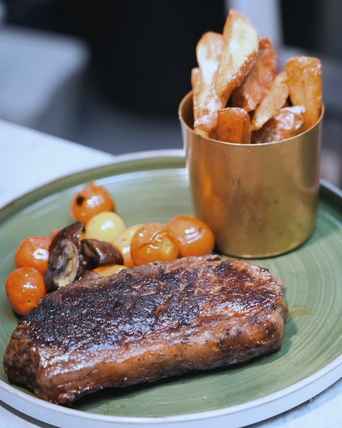 Steak on a green plate with blistered cherry tomatoes, mushrooms and a brass cup of hand-cut chips