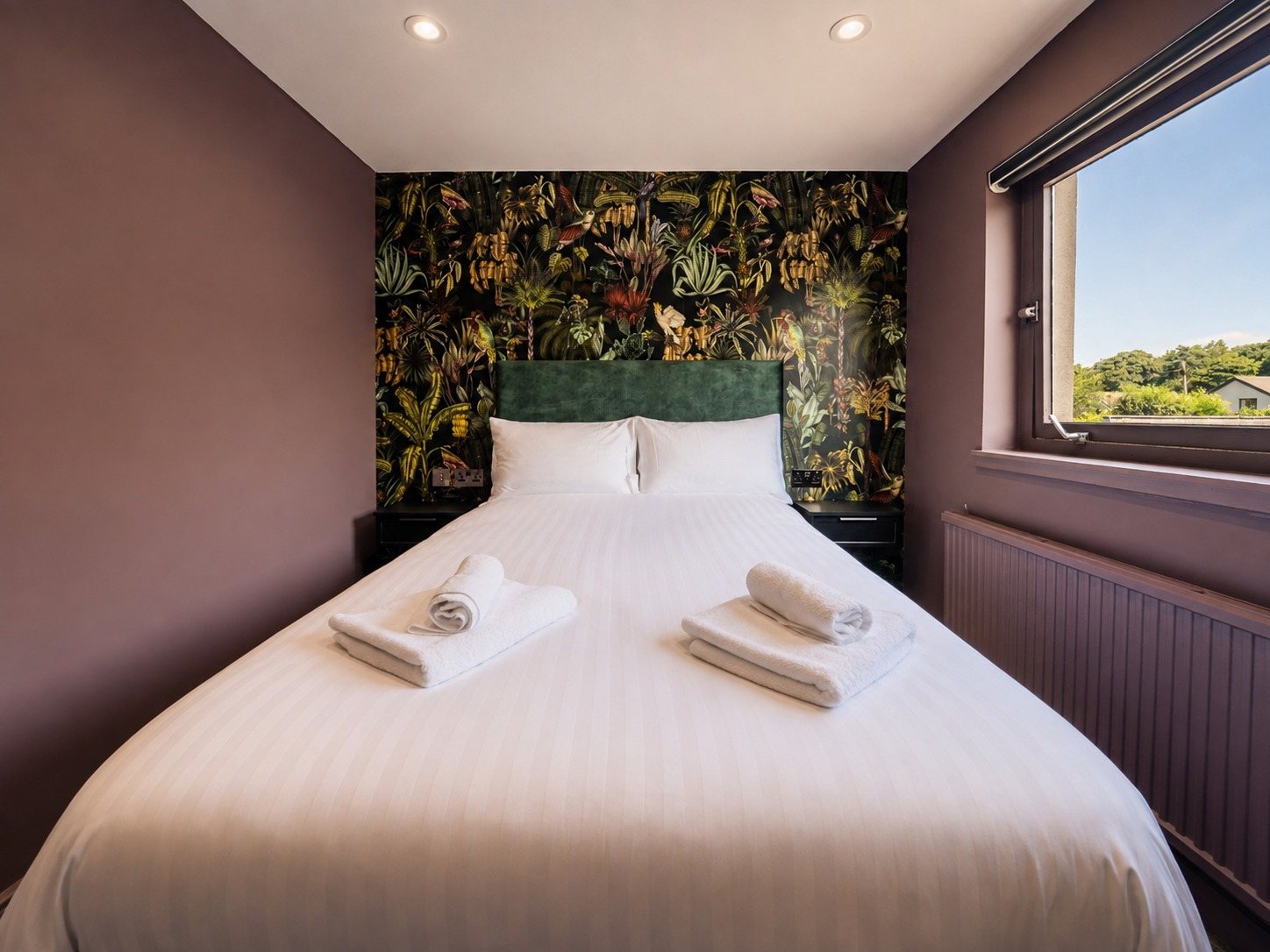 A boutique room at The Inn at Oliver Twistz with dark botanical-print wallpaper, a green velvet headboard and a made-up double bed with folded towels