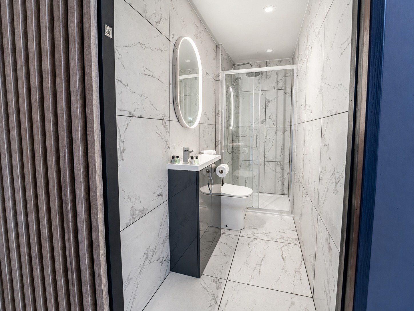 A marble-tiled en-suite bathroom with a walk-in shower, backlit mirror and vanity unit