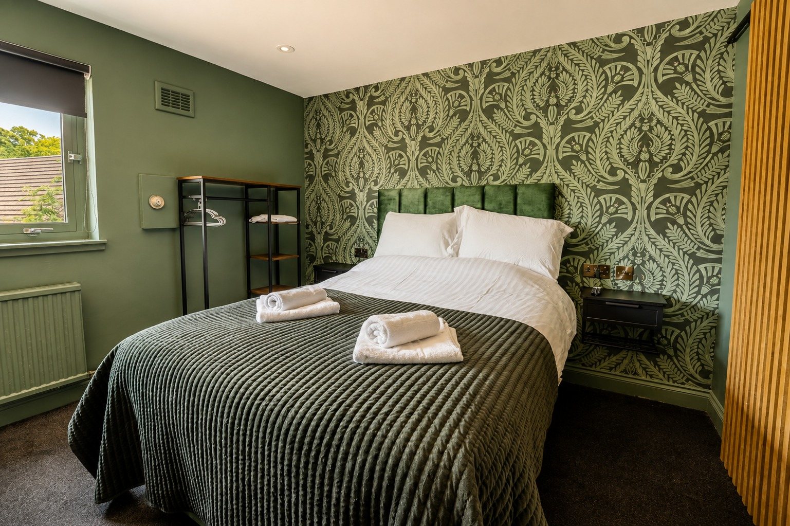 A boutique room with deep green damask wallpaper and a green velvet headboard